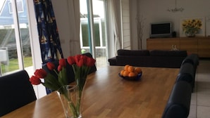 A dining area with a wooden table, a vase of red flowers, and a bowl of oranges. - Bungalow for 6 People Near Beach (Sint Maartensvlotbrug)
