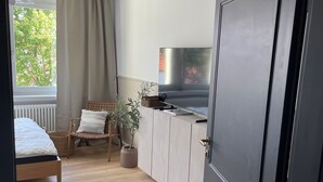 Une chambre à coucher moderne avec un lit, un plancher en bois, une fenêtre avec des rideaux, un téléviseur et une porte.