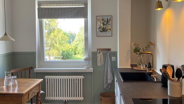 Une cuisine avec une fenêtre, un radiateur, une table avec des chaises, un tapis et un évier avec des ustensiles.
