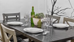Una mesa de comedor dispuesta con platos, cubiertos y cristalería, flanqueada por sillas, con una planta decorativa y una botella verde sobre la mesa.