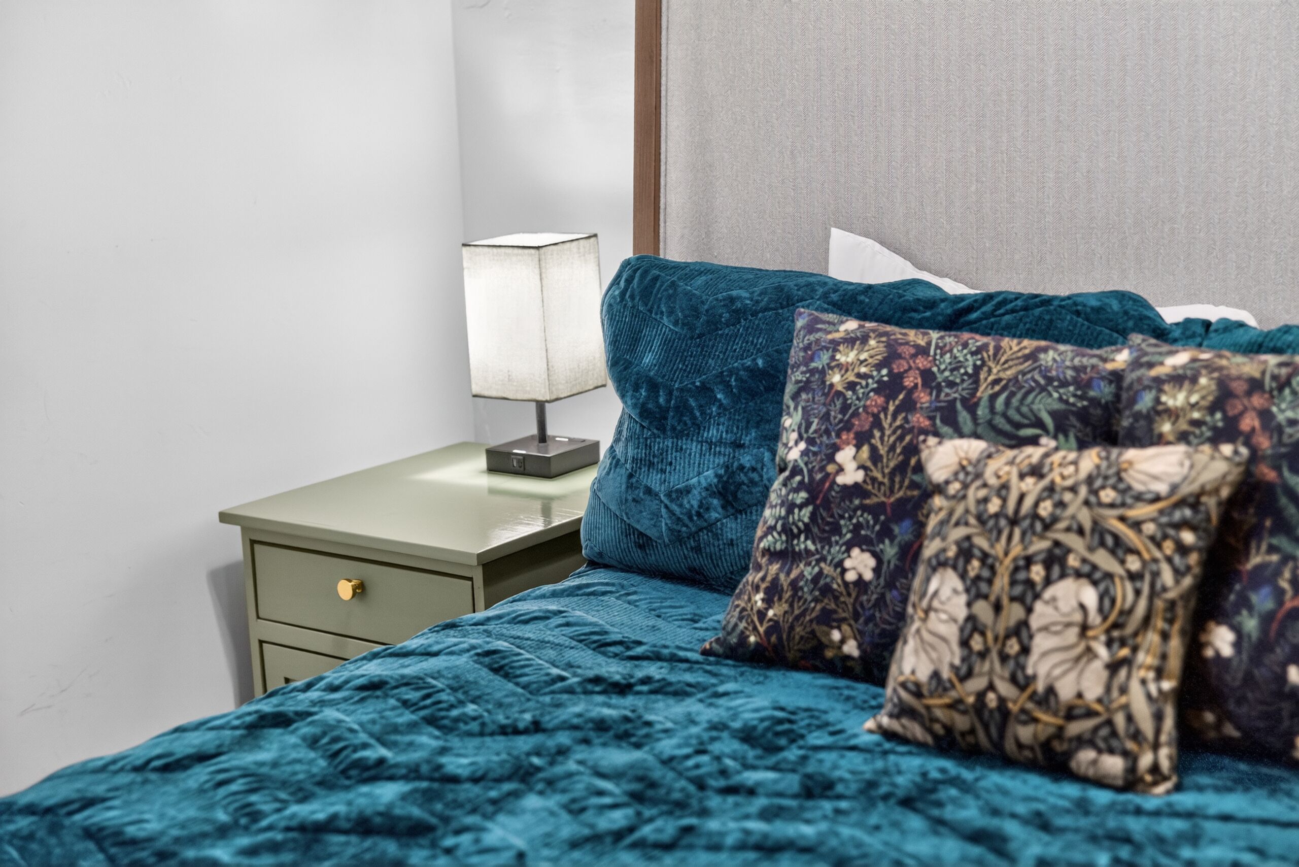Un lit avec une couette turquoise et des oreillers à motifs, une table de chevet avec une lampe, et une petite table de nuit verte avec un tiroir.
