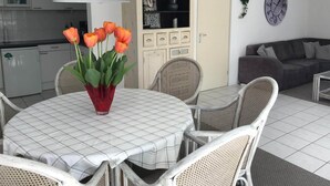 A dining area with a round table, white chairs, and a red vase with orange tulips. There is a kitchen in the background with a grey range hood and a white cabinet with decorative items. - Comfortable 6-person Holiday Home (Sint Maartensvlotbrug)