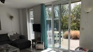 A living room with a sofa, coffee table, and a TV. There is a sliding glass door leading to an outdoor area with a table and chairs. - Comfortable 6-person Holiday Home (Sint Maartensvlotbrug)