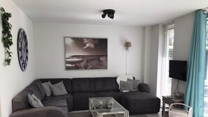A modern living room with a sectional sofa, a coffee table, a painting, a TV, and a window with curtains. - Comfortable 6-person Holiday Home (Sint Maartensvlotbrug)