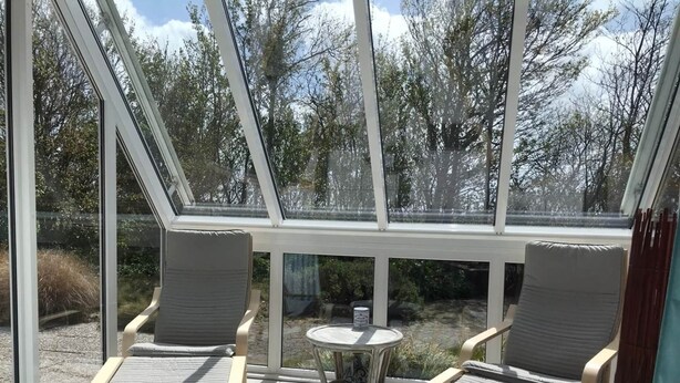 A sunlit patio with two cushioned chairs and a small table, surrounded by large windows offering a view of trees and a garden. - Comfortable 6-person Holiday Home (Sint Maartensvlotbrug)