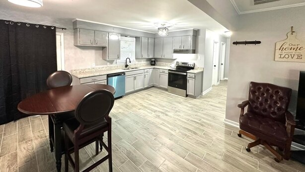 A modern kitchen with grey cabinets, a dishwasher, and a stainless steel oven. There is a dining area with a wooden table and chairs, and a leather armchair.