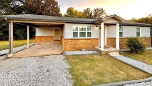 A two-story house with a covered porch, a brick foundation, and a gravel driveway.
