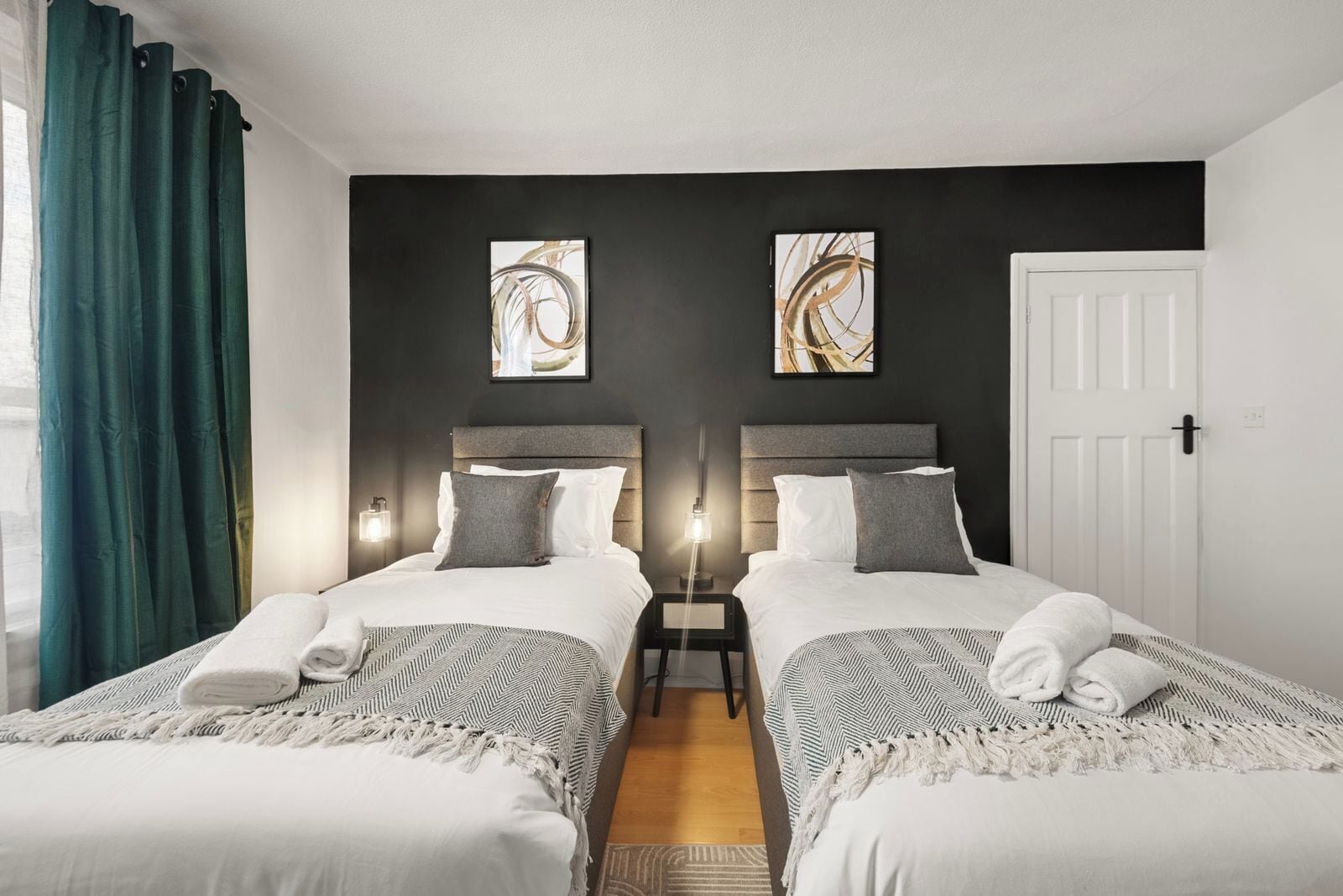 Quarto de hotel com duas camas, uma mesa de cabeceira, uma porta e obras de arte nas paredes.