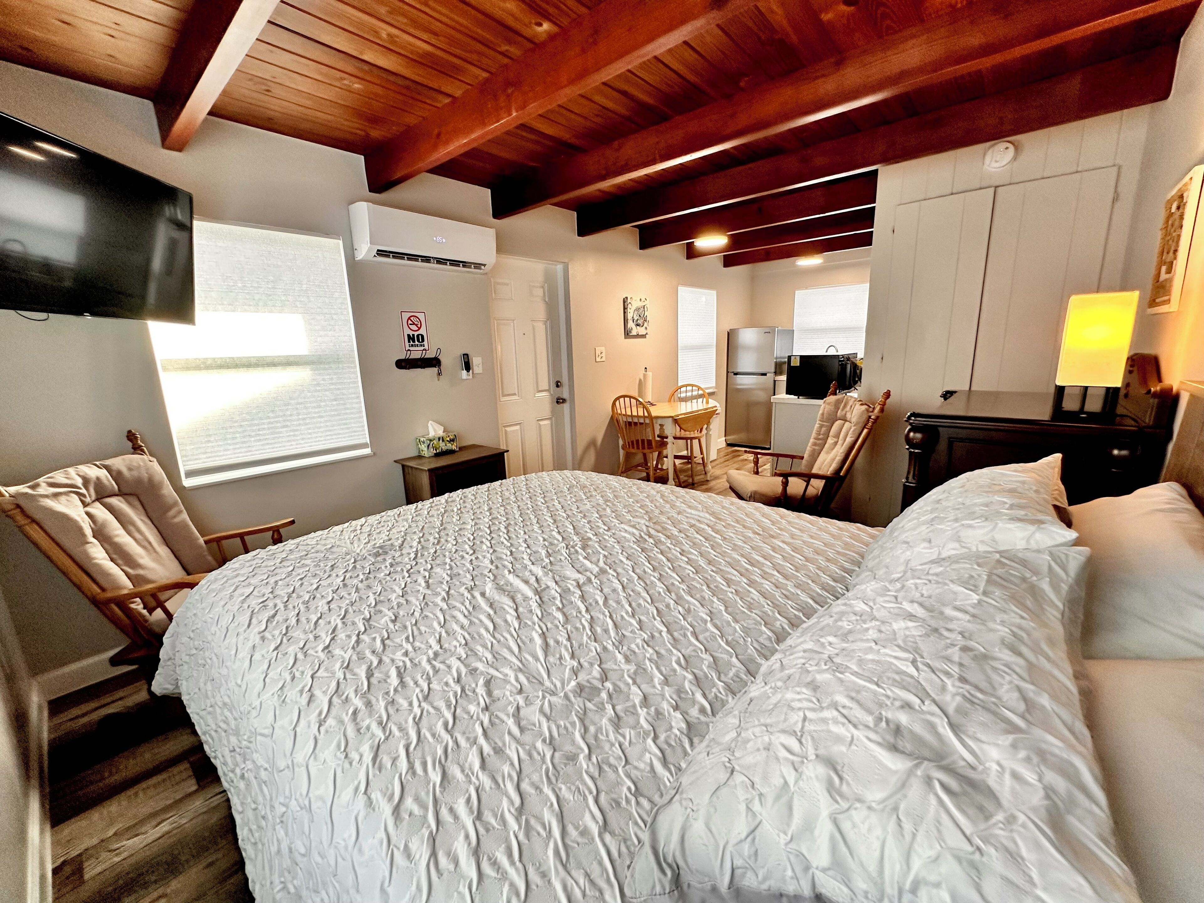 Une chambre avec un grand lit, un plafond en bois, un bureau avec une chaise, une petite kitchenette et un téléviseur fixé au mur.