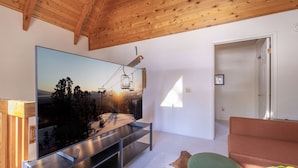 Un salon moderne avec un grand téléviseur à écran plat fixé sur un support métallique, un plafond en bois, un fauteuil vert rembourré et un canapé en cuir marron.