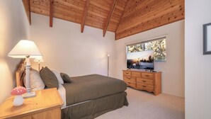 Une chambre avec un plafond en bois, un lit recouvert d’une couverture verte, une table de chevet en bois avec une lampe, une commode et un téléviseur posé sur un meuble.