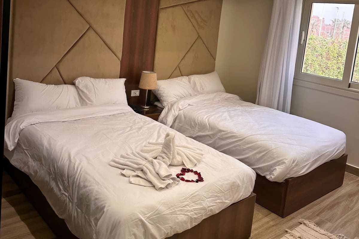 A hotel room with two beds, a large headboard, a ceiling-mounted air conditioner, a nightstand with a lamp, and a view of greenery through the window.