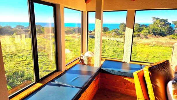Habitació amb finestrals amplis que donen vista a un camp verd i al mar. Hi ha bancs de fusta i una cadira groga.