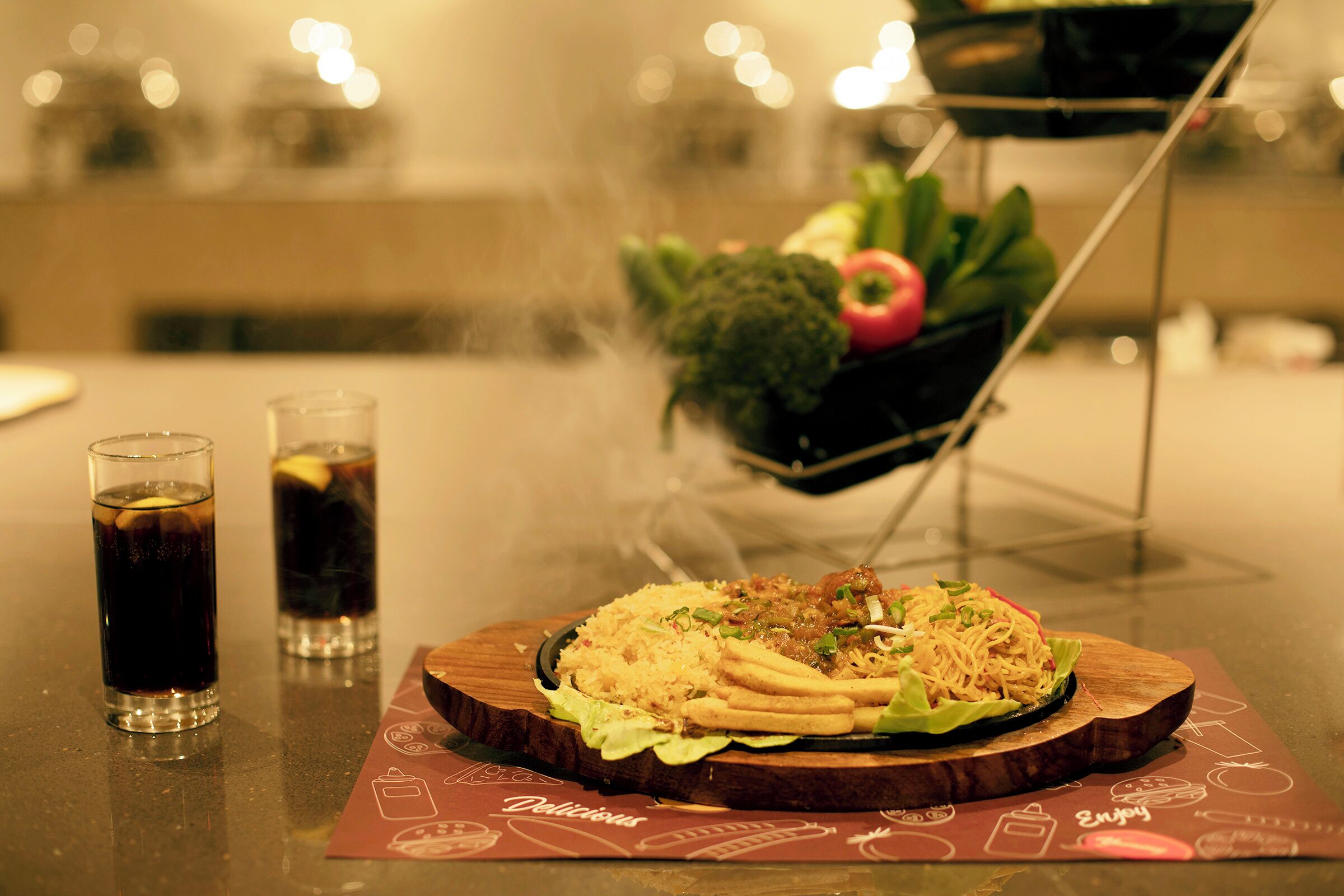 A dining table with a plate of food, two glasses of a dark beverage, and a small plant on a stand.