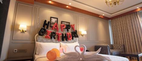 Habitación de hotel con cama, mesita de noche, lámpara, candelabro y decoración con un globo que dice 'Happy Birthday'.