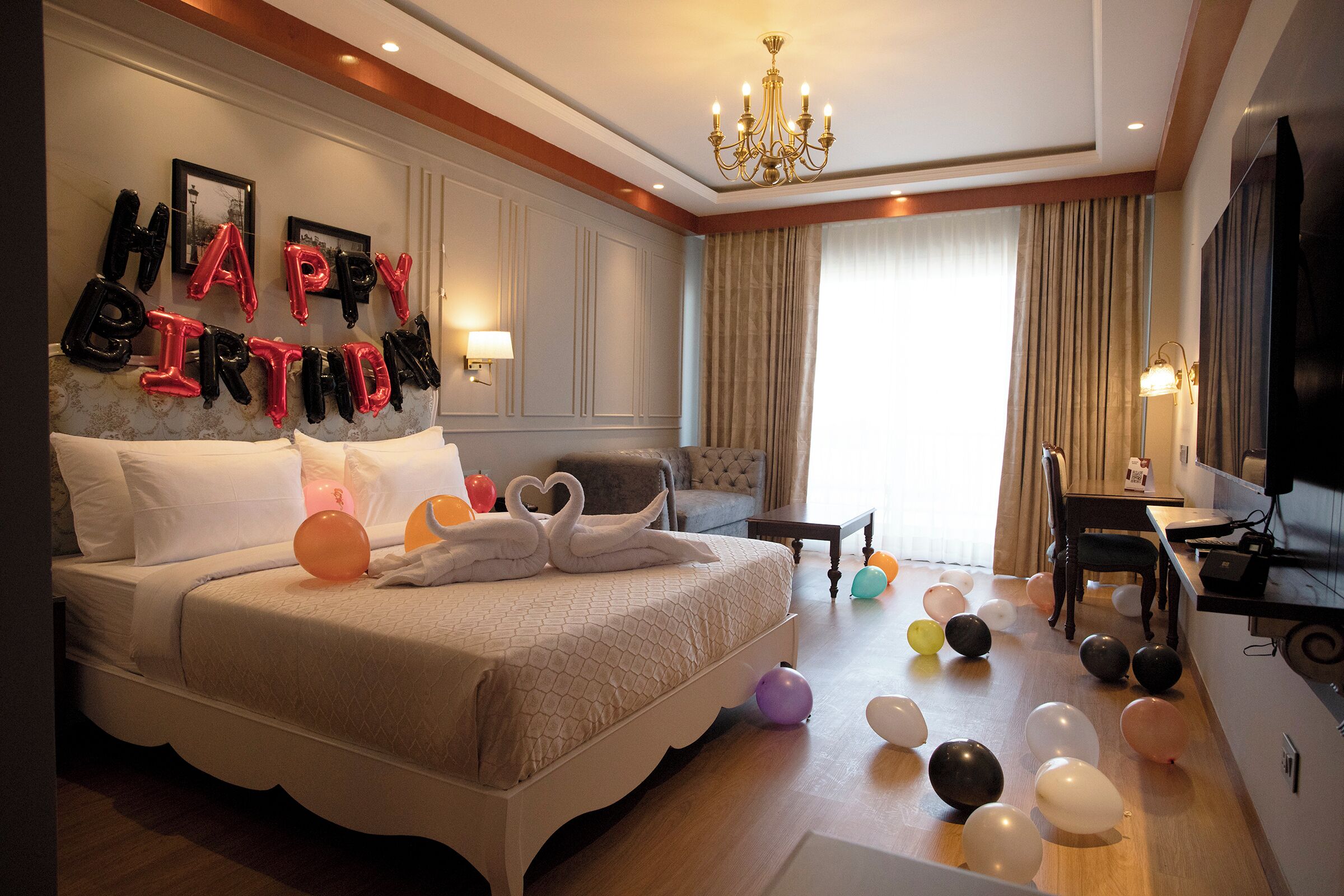 Un dormitori elegant amb un llit gran, un penjoll i una paret decorada amb globus que diuen 'HAPPY BIRTHDAY'.