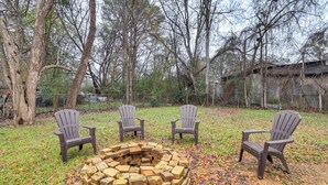 A circular brick fire pit surrounded by four Adirondack chairs in a grassy outdoor area with trees and a fence in the background. - 3 Mi to Mississippi Stadium: Cozy Jackson Getaway! (Jackson)