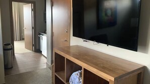 Habitación de hotel con un mueble bajo de televisión de madera, un televisor de pantalla plana montado en la pared, un cesto de basura y un pasillo que conduce a otra habitación.
