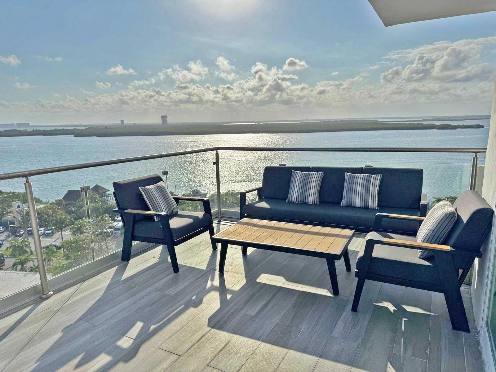 A balcony with a sofa, chairs, and a table, overlooking a body of water and distant land.