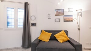 A room with a grey sofa, two yellow cushions, a window with curtains, a wall clock, and framed pictures including one of the Colosseum. - La Dolce Vita Apartment - metro Jonio (Roma)