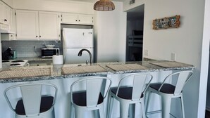 A modern kitchen with white cabinets, a stainless steel refrigerator, a built-in oven, and a bar area with white bar stools. - Modern, luxurious 1-bedroom condo in stunning Ocean Isle Beach (Ocean Isle Beach)