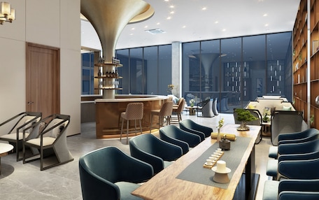 A modern dining area with a long wooden table, blue upholstered chairs, and a central bar area with shelves of bottles and glasses.