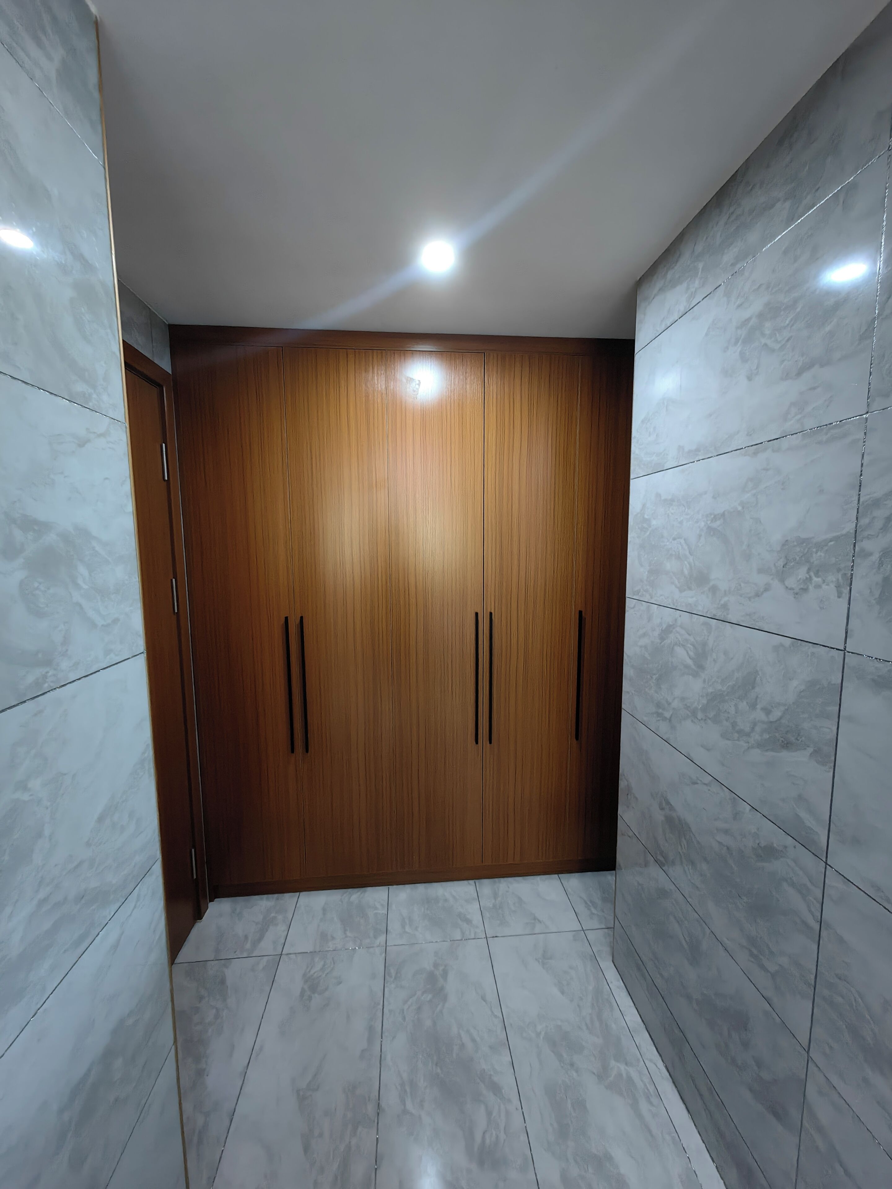 A room with a wooden wardrobe, white marble tiles, and a recessed ceiling light.