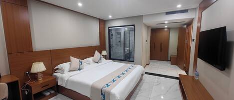 A hotel room with a large bed, wooden headboard, a TV, a desk, a lamp, and a bathroom with a glass door.