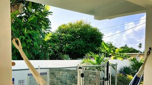 Una hamaca suspendida entre una pared blanca y una puerta de cristal, con vistas a una piscina y un patio con baldosas, y abundante vegetación al fondo.