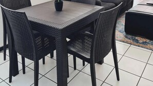 Un comedor con una mesa y sillas de mimbre oscuro, una planta en maceta y un sofá negro con cojines estampados.