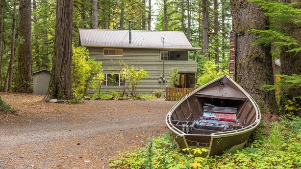 A cabin with a gravel driveway, surrounded by tall trees and a boat in the foreground. - Up-River House on the McKenzie River  (Mckenzie Bridge)