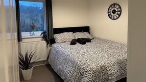 Ein Schlafzimmer mit einem Bett, einem Fenster mit Vorhängen, einer Topfpflanze, einer Wanduhr und einer Heizung.