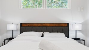 Un lit bien fait, avec du linge de lit blanc et une serviette pliée, flanqué de deux tables de chevet avec des lampes, dans une pièce dotée d’une fenêtre donnant sur des arbres.