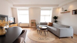 Una sala de estar moderna con un sofá gris, una mesa de centro y un comedor con mesa y sillas. Hay un televisor sobre un mueble bajo, una planta y una estantería con libros.
