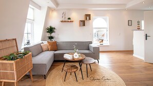 Una sala moderna con un sofá seccional gris, un sillón de ratán, una mesa de centro redonda de madera y un amplio ventanal con vistas a la ciudad.
