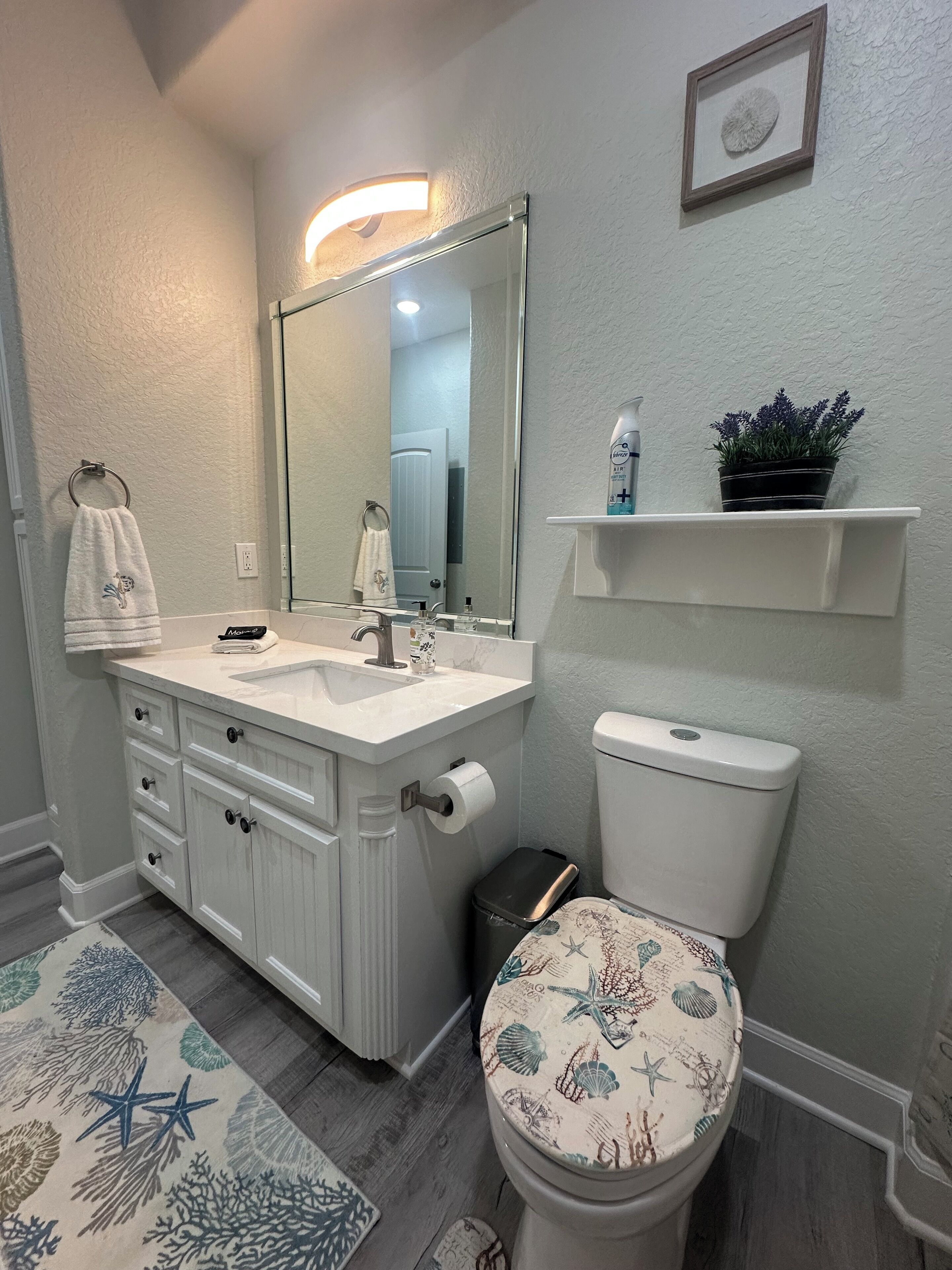 Une salle de bain avec un meuble-lavabo blanc, un grand miroir, des toilettes dotées d’un abattant décoratif, une petite poubelle et une étagère sur laquelle est posée une plante en pot.