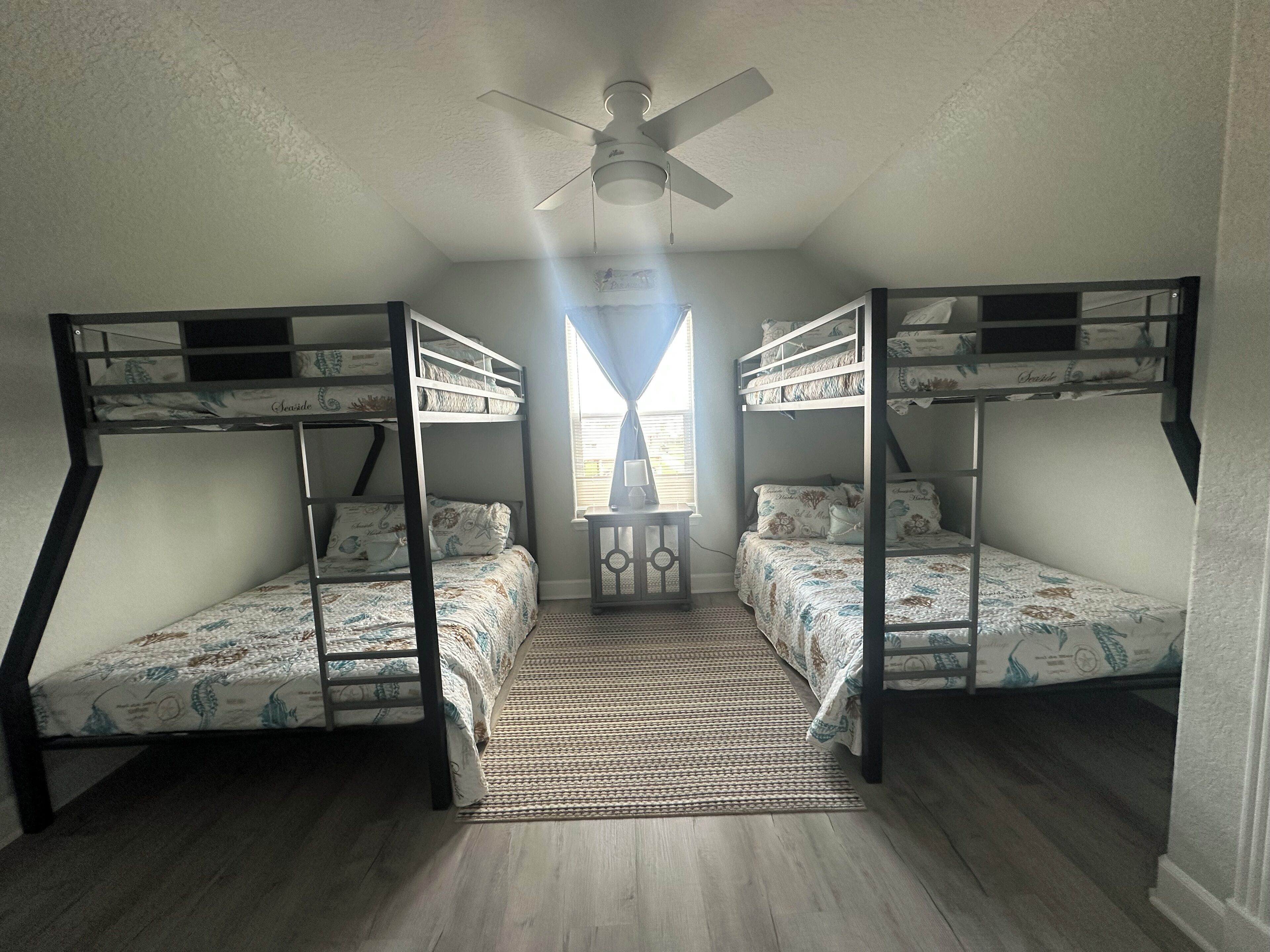 A room with two bunk beds, a ceiling fan, a window with curtains, and a small table with a lamp.