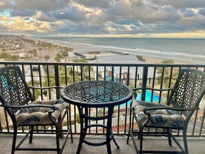 A balcony with a table and two chairs, overlooking a beach and ocean.