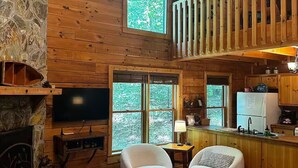 A cozy cabin interior with a stone fireplace, a large window with a view of trees, a wooden ceiling, and a kitchen area. - Misty Lake Getaway - Hot Tub/Dog Friendly/EV Charger (Blue Ridge)