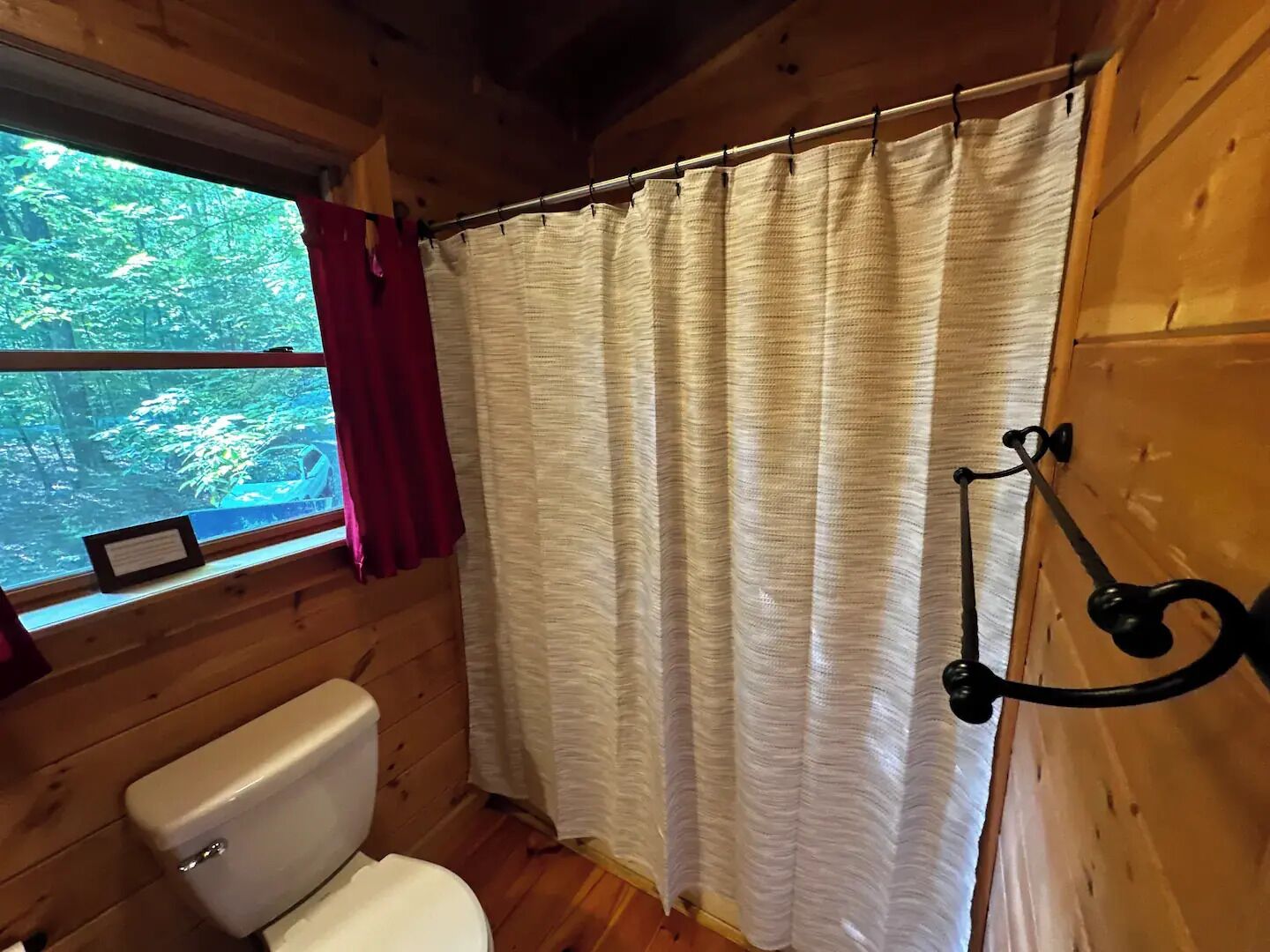 A wooden cabin bathroom with a toilet, a window with a red curtain, and a shower curtain.