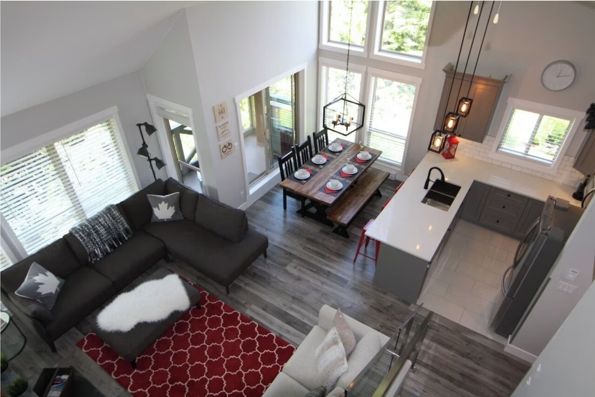 A spacious living area with a dark-colored sofa and a red patterned rug, a dining area with a wooden table and chairs, and a kitchen with a white countertop and modern lighting fixtures.