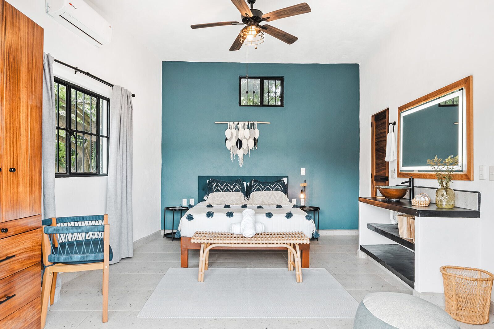 Een slaapkamer met een blauwgroene wand, een houten bedframe, een plafondventilator, een raam met tralies, een nachtkastje, een spiegel en een rieten mand.