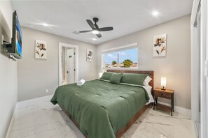 A bedroom with a bed, a ceiling fan, a nightstand with a lamp, and artwork on the walls.