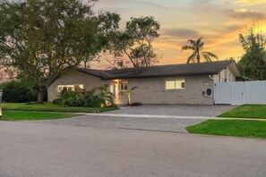A single-story house with a driveway, a front yard with trees and shrubs, and a white fence.