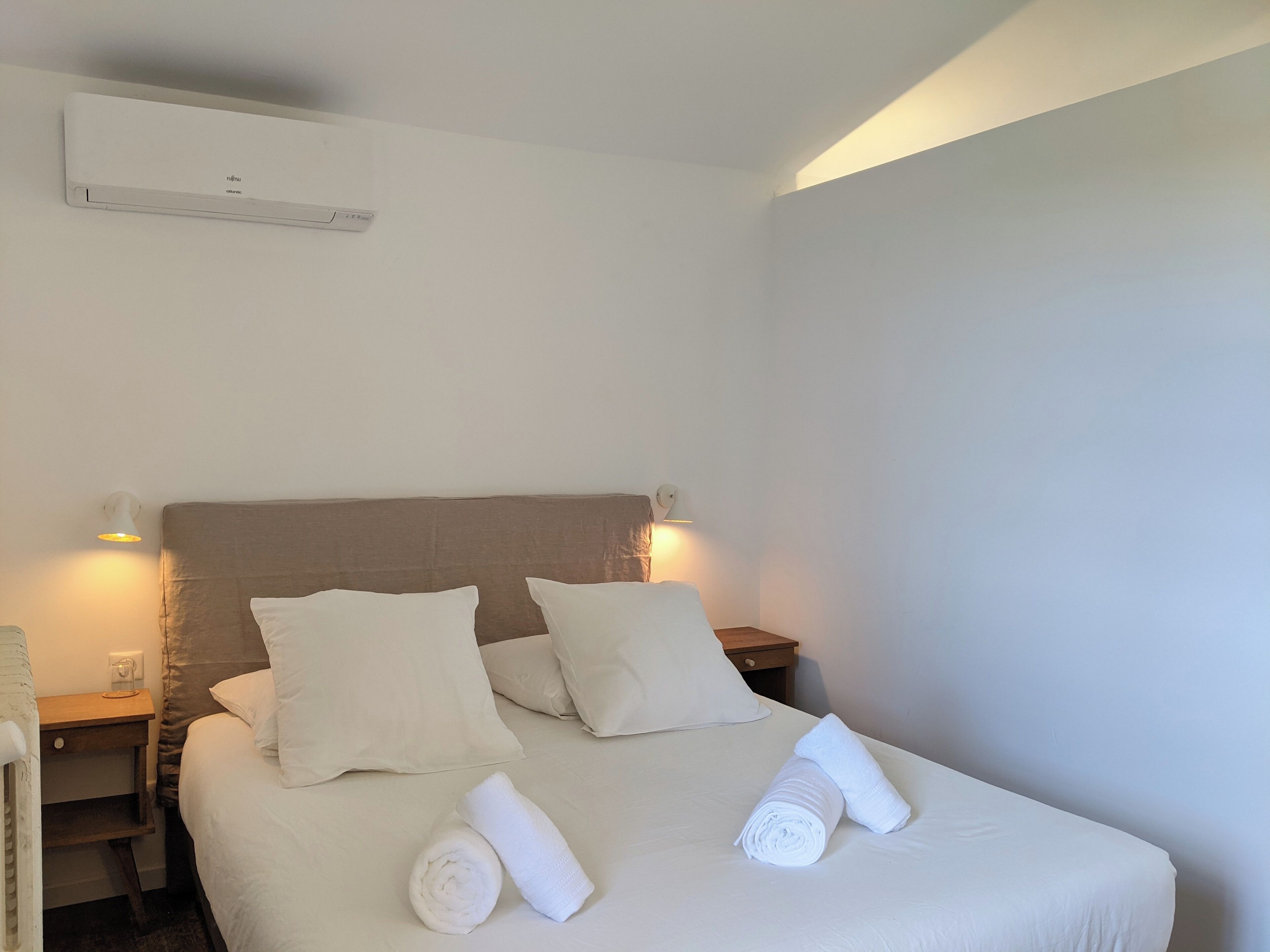 Een hotelkamer met een groot bed, twee kussens, een nachtkastje, een wandairconditioner en twee wandlampen.