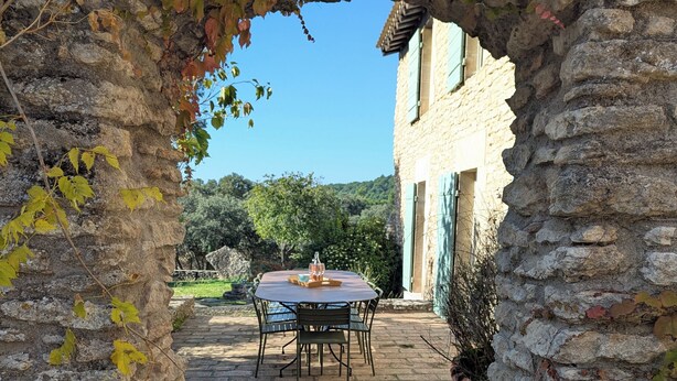 Arcada de pedra que dá para um pátio com mesa e cadeiras, rodeado por um muro de pedra e vegetação.