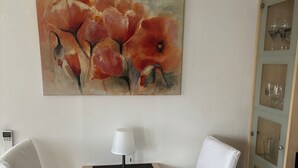 Une pièce d’angle avec un canapé blanc, une petite table en bois sur laquelle se trouve une lampe, une vitrine contenant des verres à vin et, au mur, un tableau représentant des fleurs rouges.