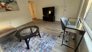 A living room with a sofa, a round table, a glass desk, a chair, a TV, and a patterned rug. - Location Lake Eola View 1 Bedroom condo (Orlando)