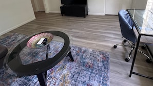 A living room with a round glass coffee table, a black TV stand, a blue office chair, and a patterned rug. - Location Lake Eola View 1 Bedroom condo (Orlando)