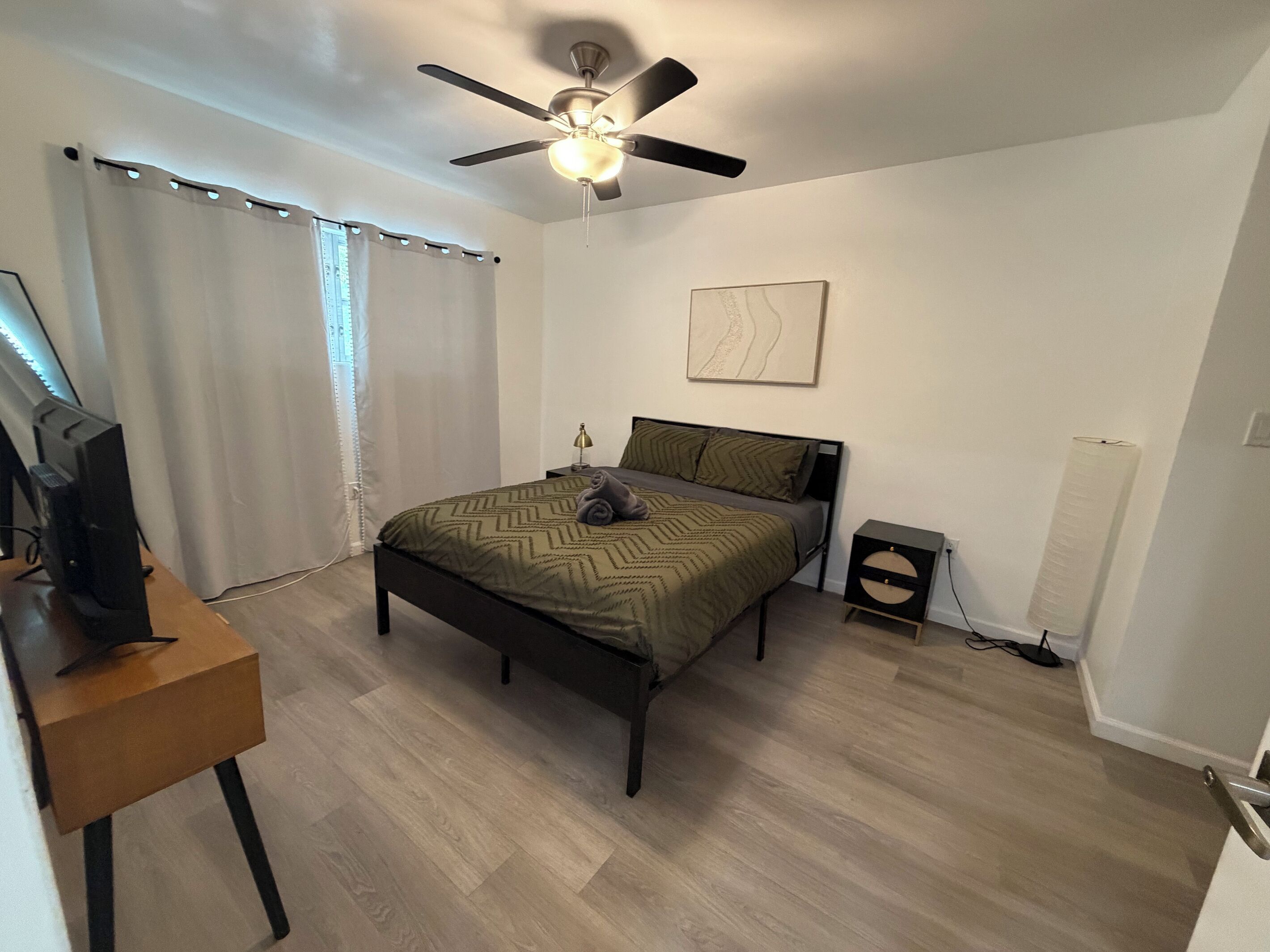 Une chambre à coucher avec un lit, un bureau avec un ordinateur, un ventilateur de plafond et une table de chevet.